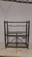 Front view of the metal folding shelf unit showing two shelves with wire mesh and folding mechanism in the center.