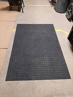 Top-down photo of a large rectangular black utility mat with a waffle grid texture, placed on a floor indoors.