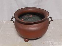 Front view showing wide flared rim, bulbous body, handle, and three rounded feet, with patina and dents visible.