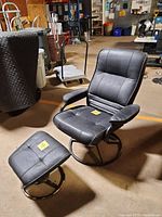 Front and angled side view of black leather look reclining chair and matching stool in indoor warehouse setting. Clear view of padded armrests, seat, backrest, and metal circular base of chair and stool.