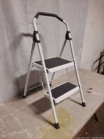 Front angle view of the white metal 2 step stool with black rubber treads, thick padded handle, and rubber feet on a worn tabletop.
