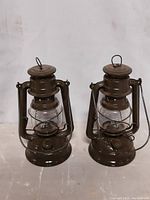 Front view of two olive green Nier Feuerhand hurricane lanterns side-by-side, showing glass chimneys and metal carriers.