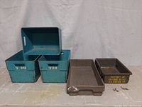 Group photo of all five storage bins: three blue metal bins stacked and two shallow grey trays to the right.