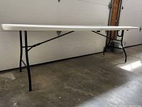Side view of the fully extended 8-foot plastic folding table with black metal legs on a concrete floor.