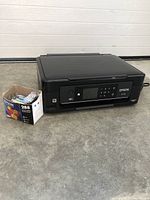 Front angled view of Epson XP-430 printer with box of new Epson 288 ink cartridges beside it sitting on concrete floor