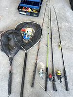 Full view of the lot showing 4 fishing rods (3 with reels), 2 fishing nets, and open tackle box with fishing gear inside.