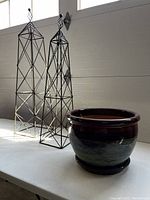 Photo showing two black metal garden obelisks of different heights alongside a ceramic garden pot with glossy multi-tone glaze on a white table.