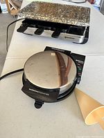 Swissmar granite top raclette maker in back, Chef's Choice waffle cone maker with cone shaper in front on a white table.