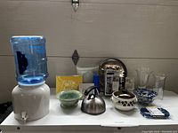 Overview of lot including ceramic water dispenser with spigot and blue jug, green and blue ceramic bowls, white crock bowl, Hamilton Beach blender box, metal tray, glass vases, yellow 'Hello Sunshine' sign, and small blue Ricardo box.
