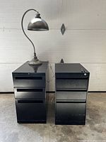 Full view of two black metal filing cabinets side by side with an industrial lamp placed on top of the smaller cabinet, showing overall size and condition.