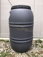 Full view of black plastic rain barrel upright on gravel ground, showing cylindrical body with horizontal ridges and a closed lid.