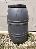 Front view of the gray plastic rain barrel showing its cylindrical shape and two bands around the body, standing on gravel with some dirt and dried grass on it.