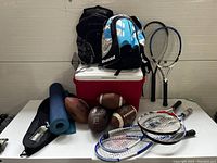 Full view of lot items including cooler, backpacks, footballs, tennis and squash racquets, yoga mat