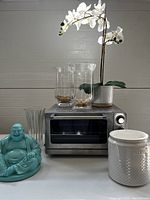 Full lot image showing toaster oven, 2 hurricane glass lanterns with pebbles, faux white orchid in ceramic pot, turquoise Buddha figurine, glass utensil holder, and white vase