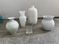 Five white and clear vases of varying shapes and sizes placed on concrete floor.