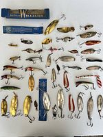 Top-down photo showing a wide spread of about 48 assorted vintage fishing lures, mostly metal with some wood, of various shapes, colors and sizes including some gold and silver shiny spoons, some painted wooden lures, and some lures still sealed in original blue Williams packaging.
