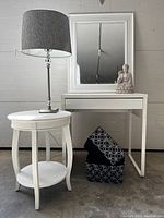 White IKEA computer desk with drawer, white framed mirror, round side table with lower shelf, modern table lamp with silver base and heather grey shade, black and white storage baskets, cream Buddha figurine arranged together.