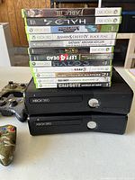 Shows two black Xbox 360 consoles stacked with a stack of eleven Xbox 360 game cases on top, and two camouflage and one black controllers placed around.