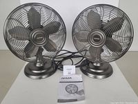 Two Noma oscillating table fans placed side by side with user manual in front, showing overall condition and size.