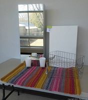 Table top displaying colorful bath mat, two white plastic cups, two wire baskets, and a white melamine cabinet leaning upright.
