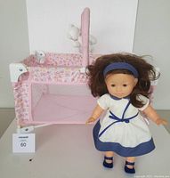 Photo showing Corolle baby doll standing beside pink floral playpen style doll crib with toy bar and two dangling toys.