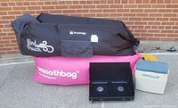 Two black and pink inflatable air loungers with their storage bags, Ventura propane camp stove, and Coleman personal cooler together on paved surface in front of brick wall.