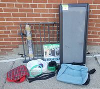 View of multiple dog travel items including PetSafe car ramp, pet barriers/gates, blue Kurgo dog carrier, travel bowl, dog coat, collar, and leash arranged outside against brick wall.