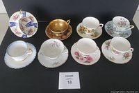 Lot of 8 assorted vintage cups and saucers on black background with lot number card. Various floral designs and gold trim visible.