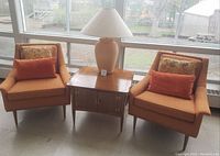 Front view of two orange armchairs flanking wooden storage table with decorative cushions on chairs