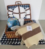 Wicker picnic basket with arranged picnic items including two plates, four mugs, plastic cutlery, blue and white checkered tablecloth, beige and brown wool blanket, and sealed Polaroid camera package.