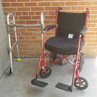 Side view of the red framed wheelchair and silver foldable walker against a brick wall.