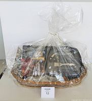 Full view of gift basket wrapped in clear plastic wrap showing contents including multiple wine accessories and glassware inside a wicker basket.