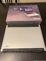 Photo shows one silver Acer laptop closed with rear ports visible, placed on a table next to a box of ASUS Transformer Book Flip laptop.