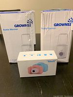 Two white boxes containing Grownsy bottle warmers and a packaged box with two children's digital cameras on a dark surface.