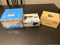 Three boxed cameras/camcorders on table; boxes visibly damaged, showing front and sides of packaging.