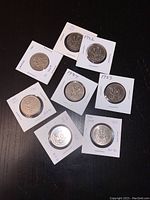Photo of eight Canadian 50 cent coins in white paper holders with year labels (1980, 1981, 1982, 1983, 1984, 1985, 1986, 1989). Coins show wear and weathered surfaces.