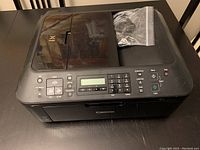 Canon MX410 printer showing top and control panel with buttons, LCD display, and plastic bag containing accessories.