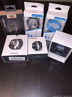 Photo of 5 unopened items: 2 smart bracelets in blue and gray, 2 boxed sports watches (WATCH1 and Lebistar), 1 black Escape TWS earphones with charging case
