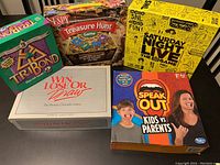 Lot of six colorful board and party game boxes arranged on table: TriBond, I Spy Treasure Hunt, Saturday Night Live The Game, Win Lose or Draw 1st Edition, and Speak Out Kids vs Parents