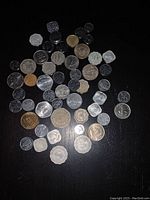 Photo showing 52 assorted foreign coins of various sizes, shapes, and metals laid out on a black surface. Coins show wear and weathering, some have scalloped edges, others round. Designs include numeric denominations, script, and various emblems.