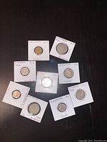 Nine foreign coins displayed individually in white cardboard holders with handwritten country and year labels, arranged on a dark surface.