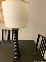 Table lamp with a black ribbed base and beige drum lamp shade placed on a black dining table with black chairs in the background.