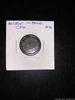 Front side of ancient coin in protective white cardboard holder with visible weathering and corrosion.