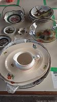 Varied china plates including larger floral designs, cup and saucer, glass gold-rimmed plate with handle, and small decorative plates with scenic and cultural artwork.