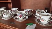 View of six floral porcelain teacups and saucers on wooden shelf, showing details of floral patterns and gold trim.