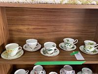 Wide shot showing five cups and saucers lined up on a wooden shelf in a single row, displaying their distinct floral patterns.