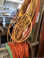 Three outdoor extension cables: one orange cable coiled on wooden reel, two longer yellow and orange cables hanging loosely on hooks.