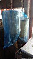 Two hanging lights with beige cylindrical fabric lampshades hanging, one covered with blue plastic wrapping, viewed near window and counter.