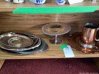 Photo showing stack of metal trays on shelf, copper mug with tray, and decorative silver plate on a pedestal stand.
