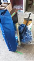 Blue folding chair in blue carrying bag next to a rolled tarp and other equipment on concrete floor